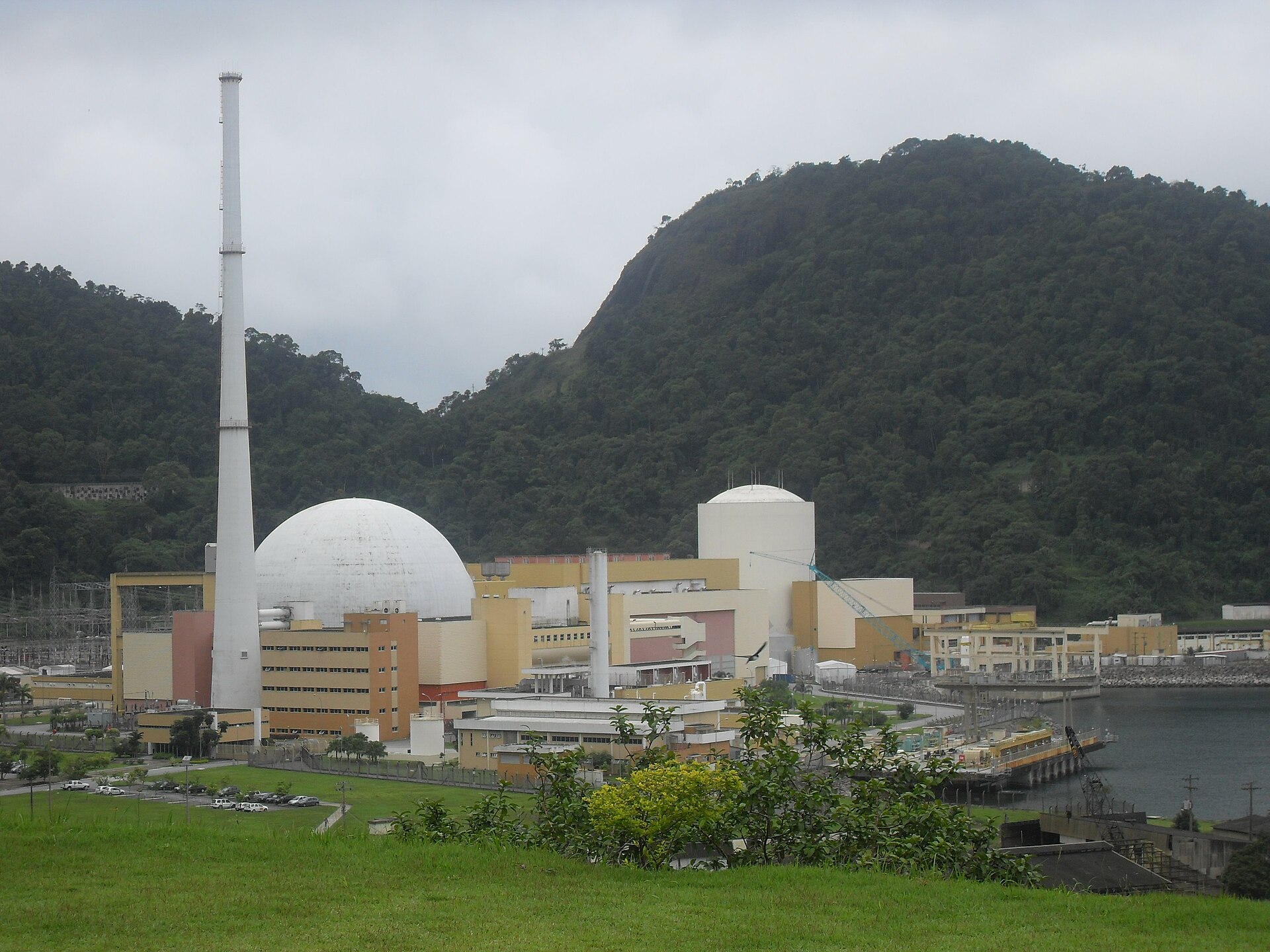 Nuclear power plant buildings with a white dome and tall chimney below a forested hill at Angra dos Reis, under an overcast sky