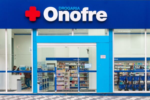 The blue and white front of a Brazilian pharmacy with a red cross logo, an open entrance and shelves of products inside