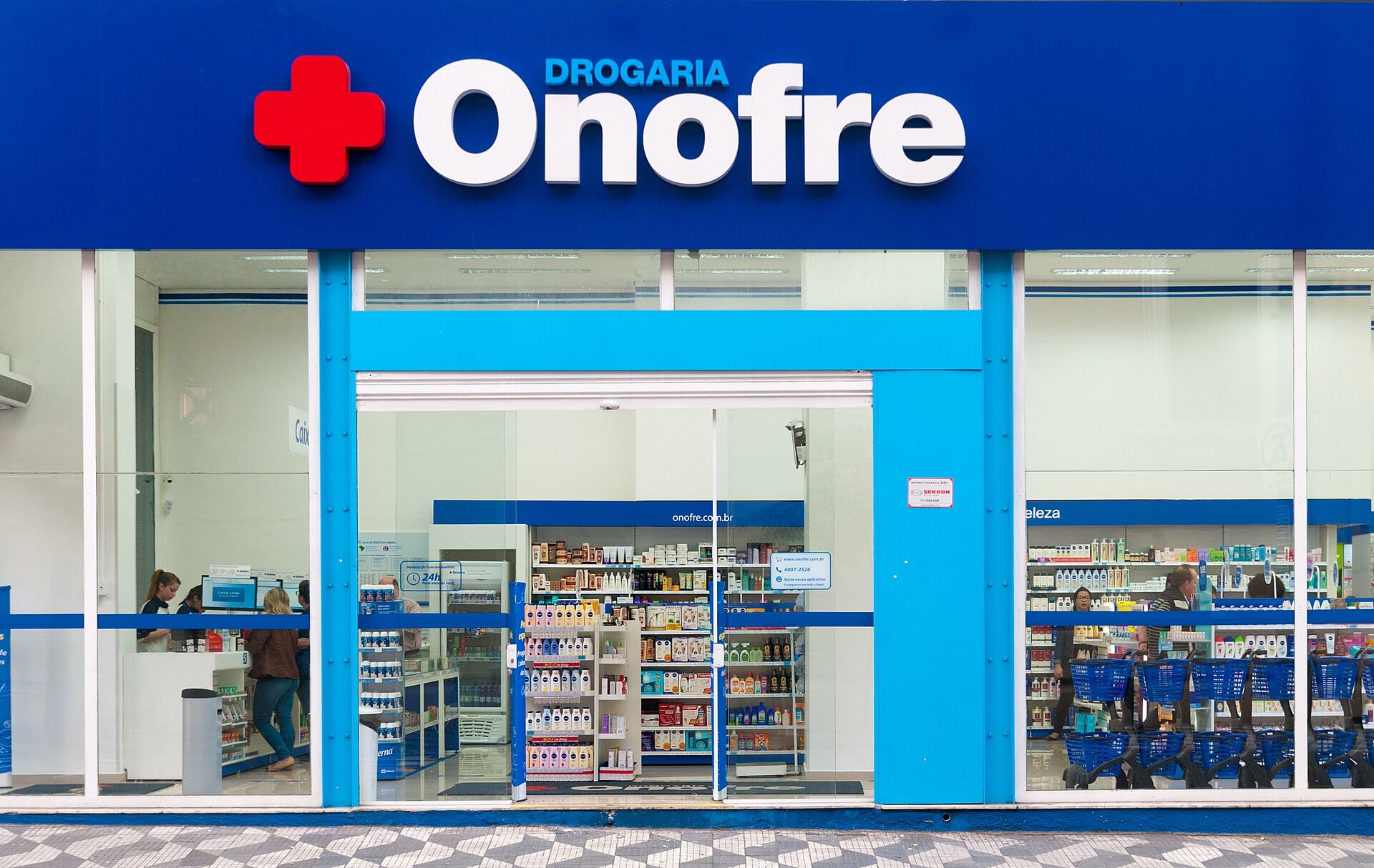 The blue and white front of a Brazilian pharmacy with a red cross logo, an open entrance and shelves of products inside