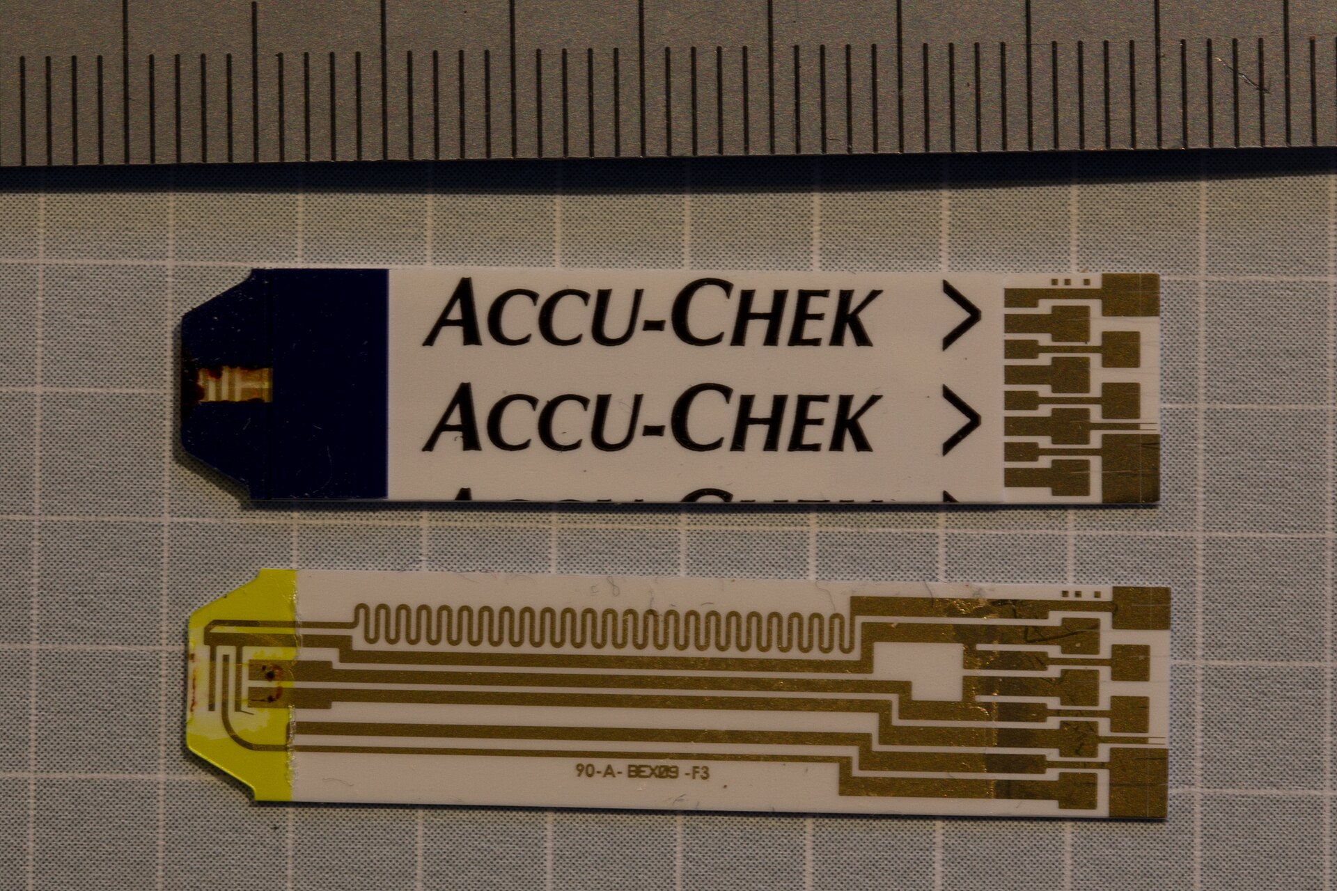 Two Accu-Chek blood glucose test strips on a grid mat beside a ruler, the lower one with its cover peeled off to show the circuit