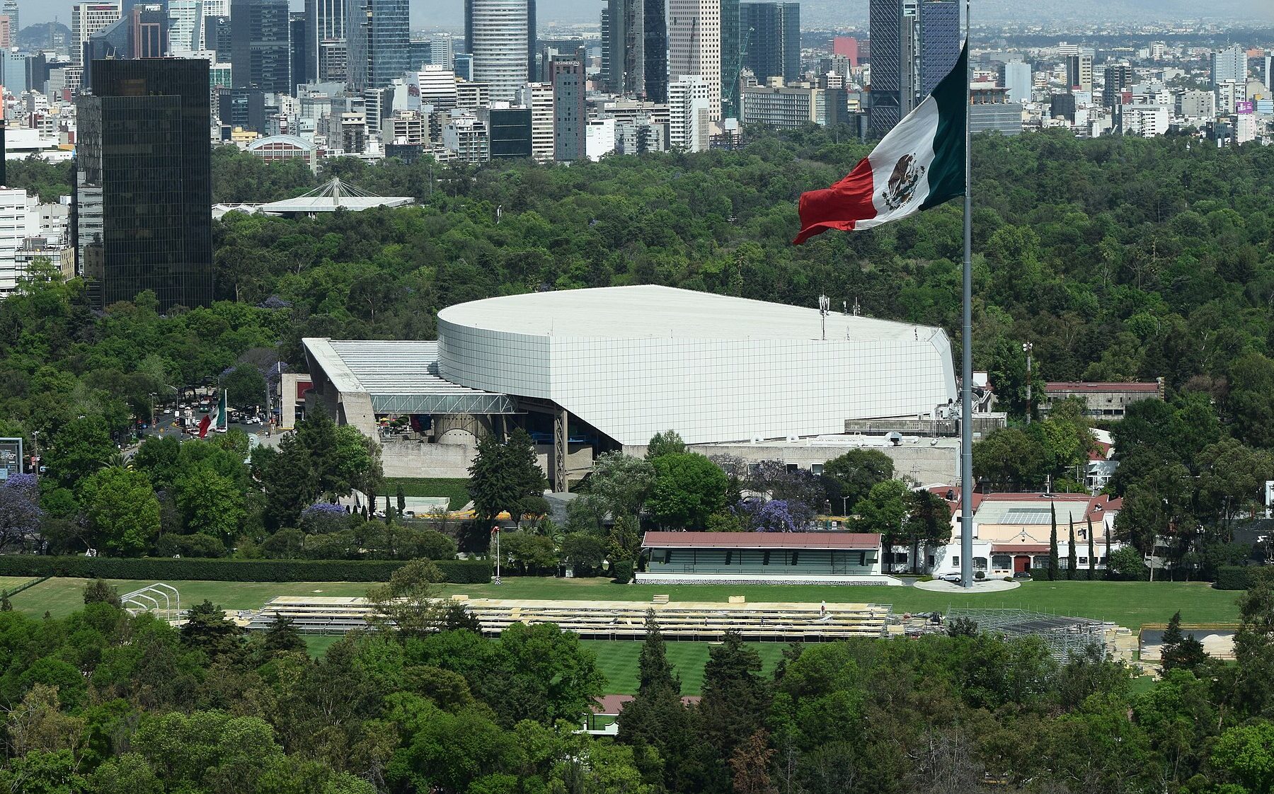 A giant Mexican flag on a tall pole above green parkland and a white curved-roof building, with Mexico City towers on the skyline