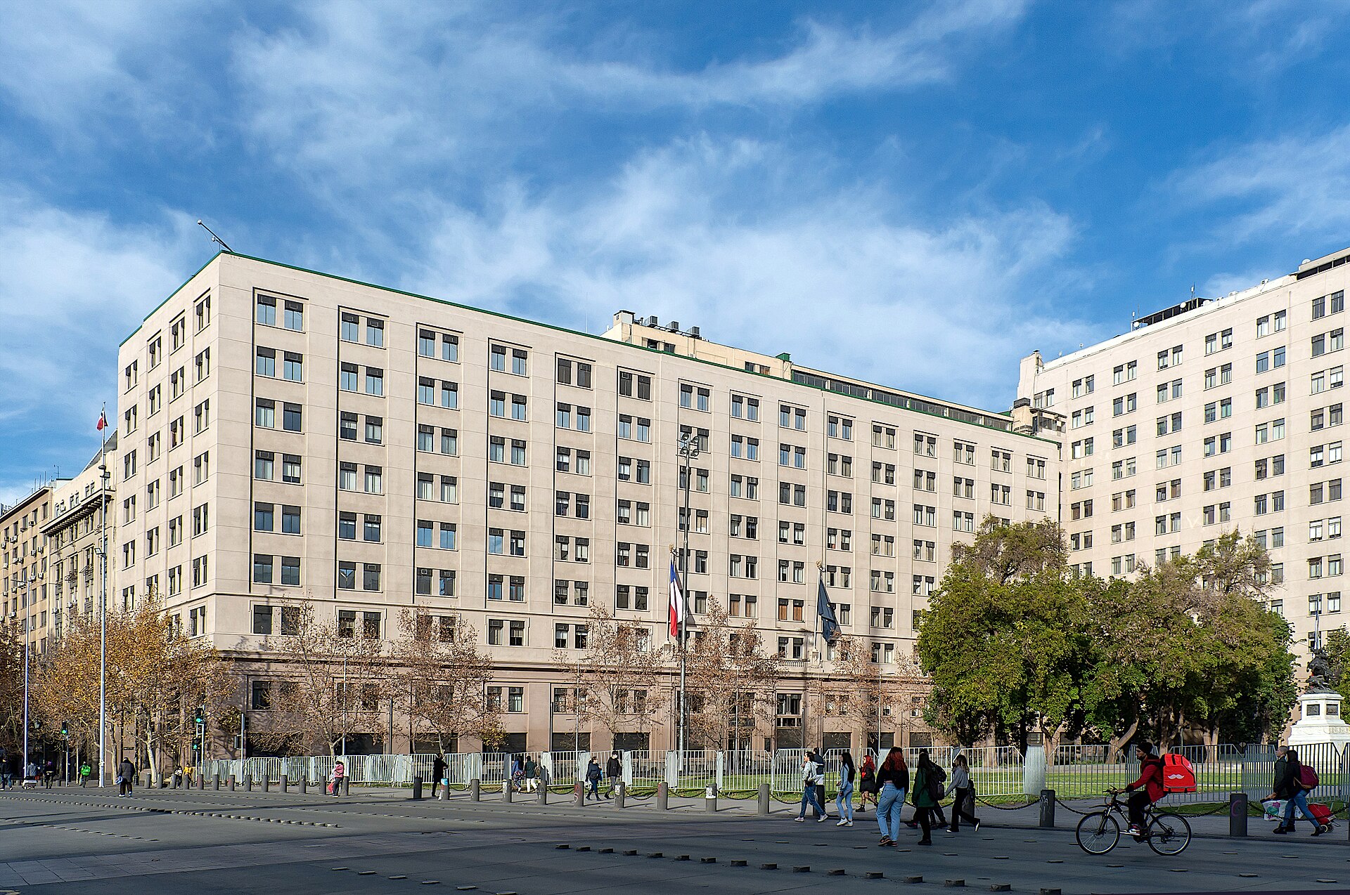 A large cream-coloured government office building in central Santiago under a blue sky, with trees, flagpoles and pedestrians in front