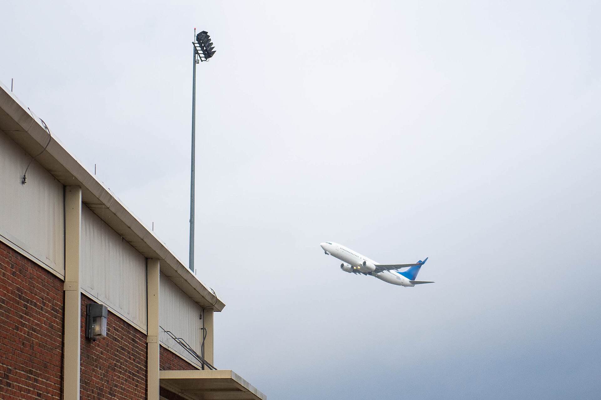 A white charter jet with a blue tail climbing into an overcast sky beside a brick building and a floodlight mast