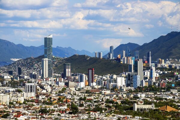 High-rise towers of Monterrey rising above low-rise neighbourhoods, with green hills and the Sierra Madre mountains under a cloudy blue sky