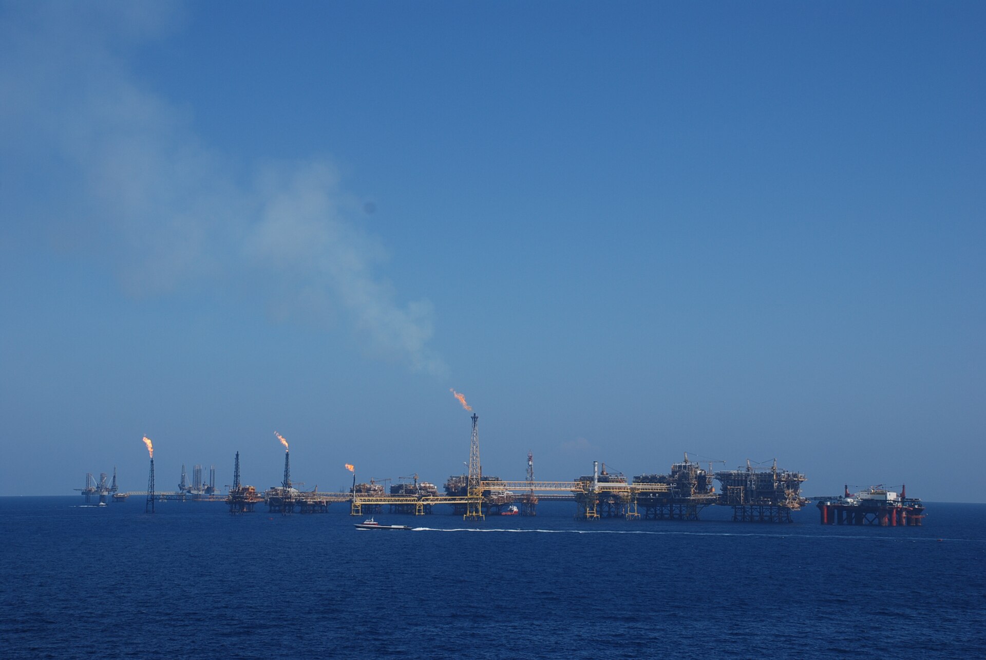 Pemex offshore oil platforms linked by bridges on a calm blue sea, with gas flares burning and a supply boat passing in front