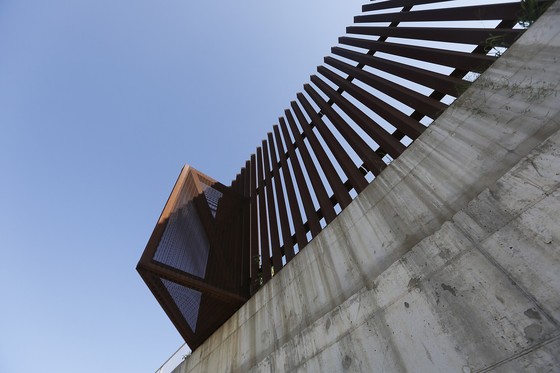 Rust-coloured steel bollards rising above a sloping concrete wall against a blue sky, seen from below