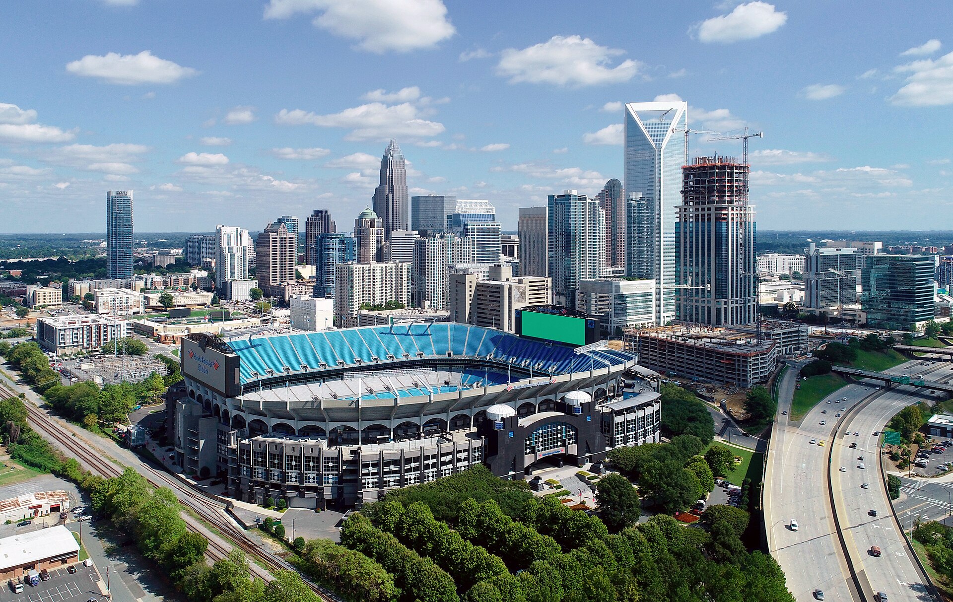 Aerial view of downtown Charlotte with skyscrapers, a blue-seated stadium in the foreground and a highway on the right under a blue sky