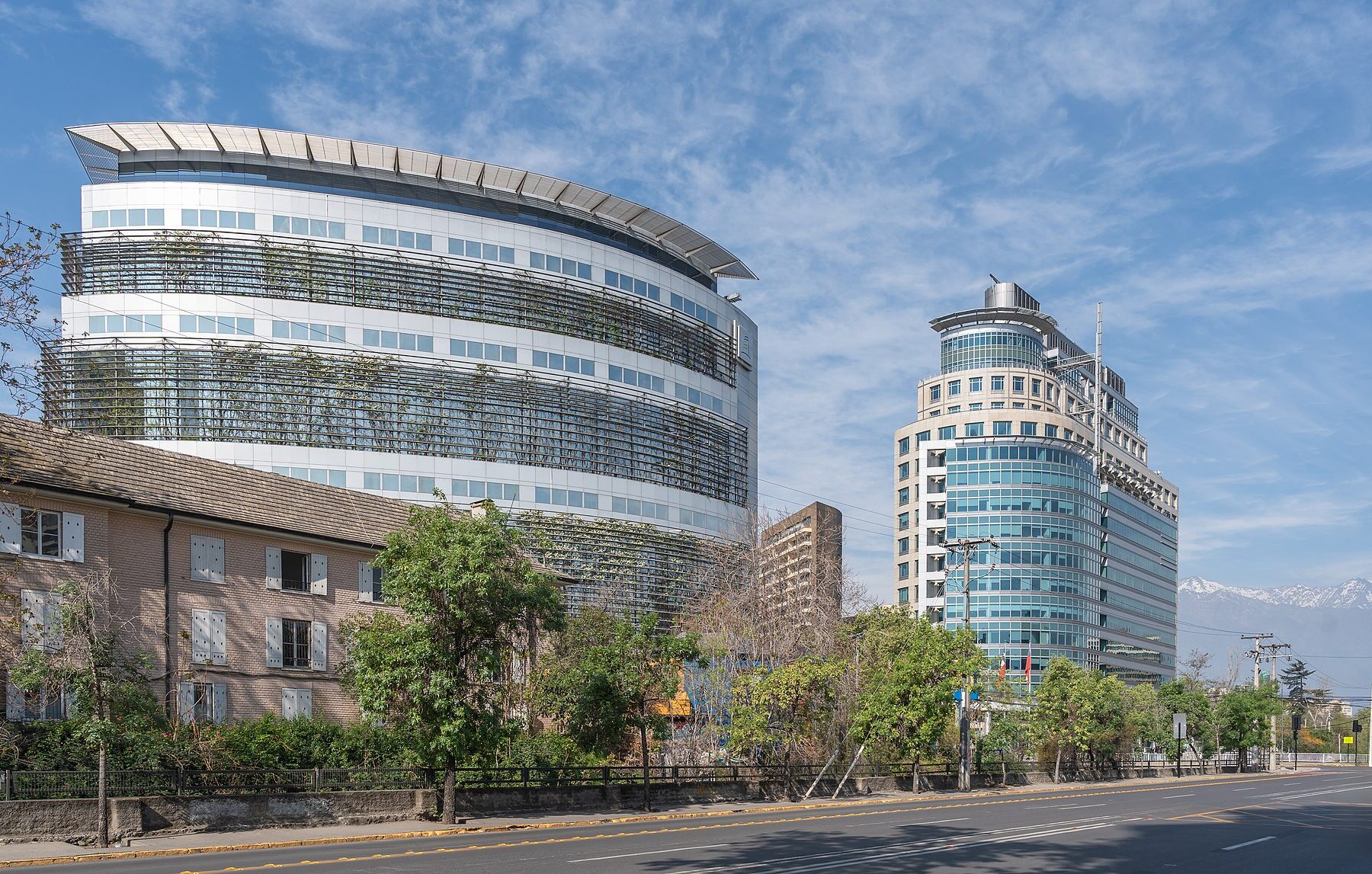 Two modern office towers, one curved with a green facade screen, beside a tree-lined street in Santiago with snow-capped Andes peaks at right