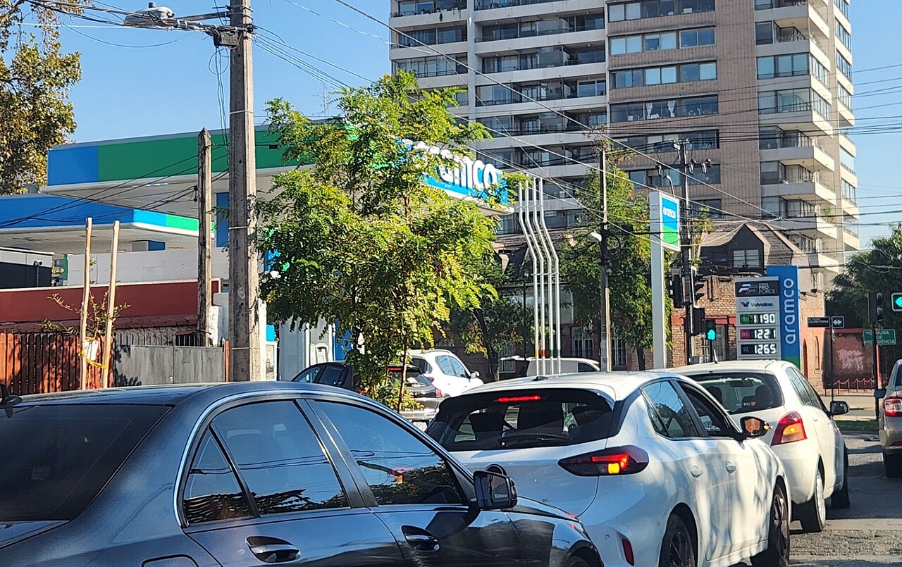 Cars queue on a Santiago street beside a green-and-blue Aramco petrol station, with apartment blocks and trees behind