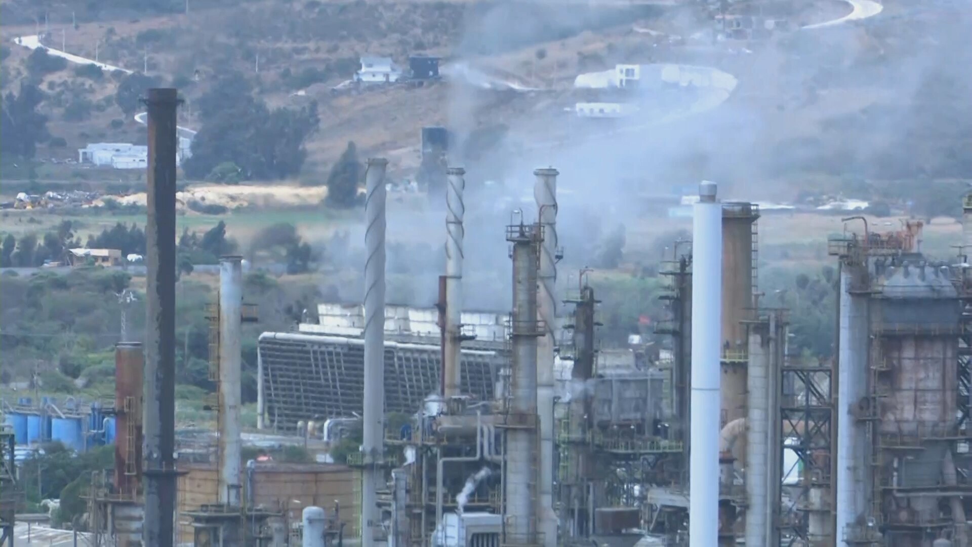 Chimneys, steam and processing units at an oil refinery on a hillside, with a cooling structure in the middle