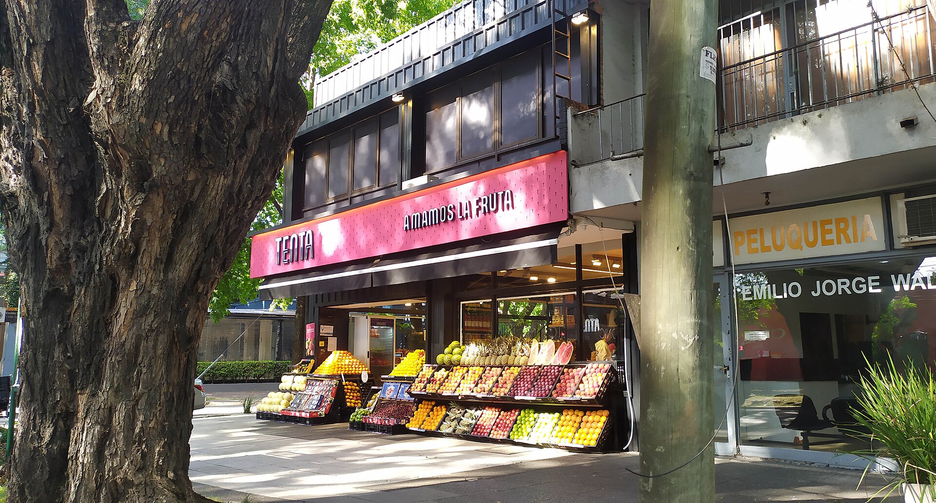 A greengrocer's shopfront with a pink sign and crates of oranges, apples and other fruit stacked on the pavement under a tree in Martinez, Buenos Aires province