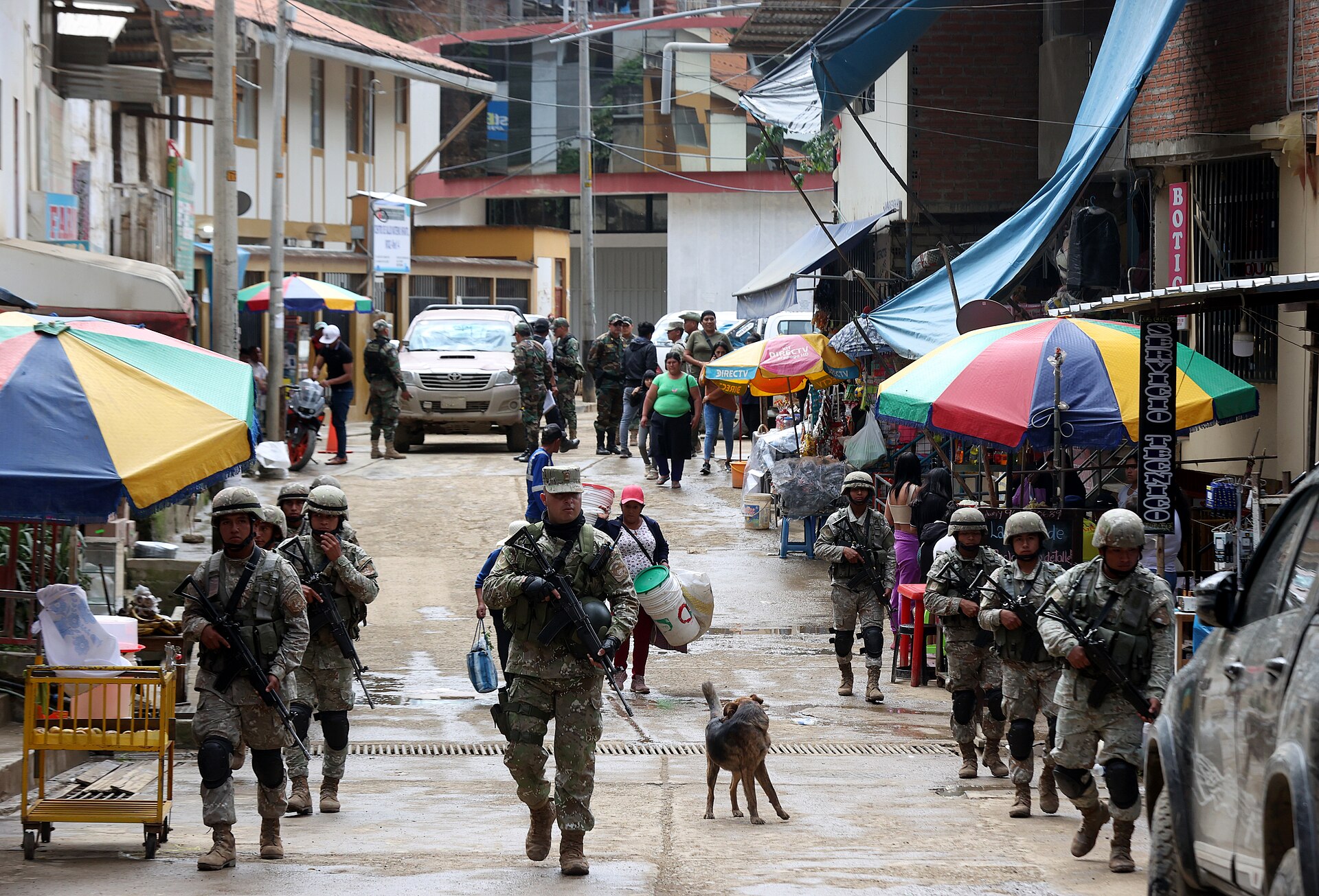 Soldiers in camouflage and helmets with rifles patrol a wet market street in Pataz past umbrellas, stalls and a pickup truck
