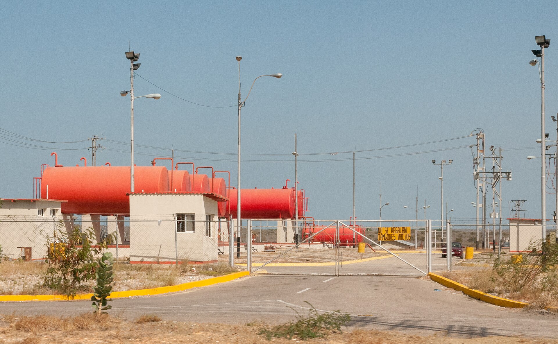 Red fuel tanks, floodlight poles and a gated entrance at a Corpoelec gas thermoelectric station on Margarita Island, Venezuela under a clear sky