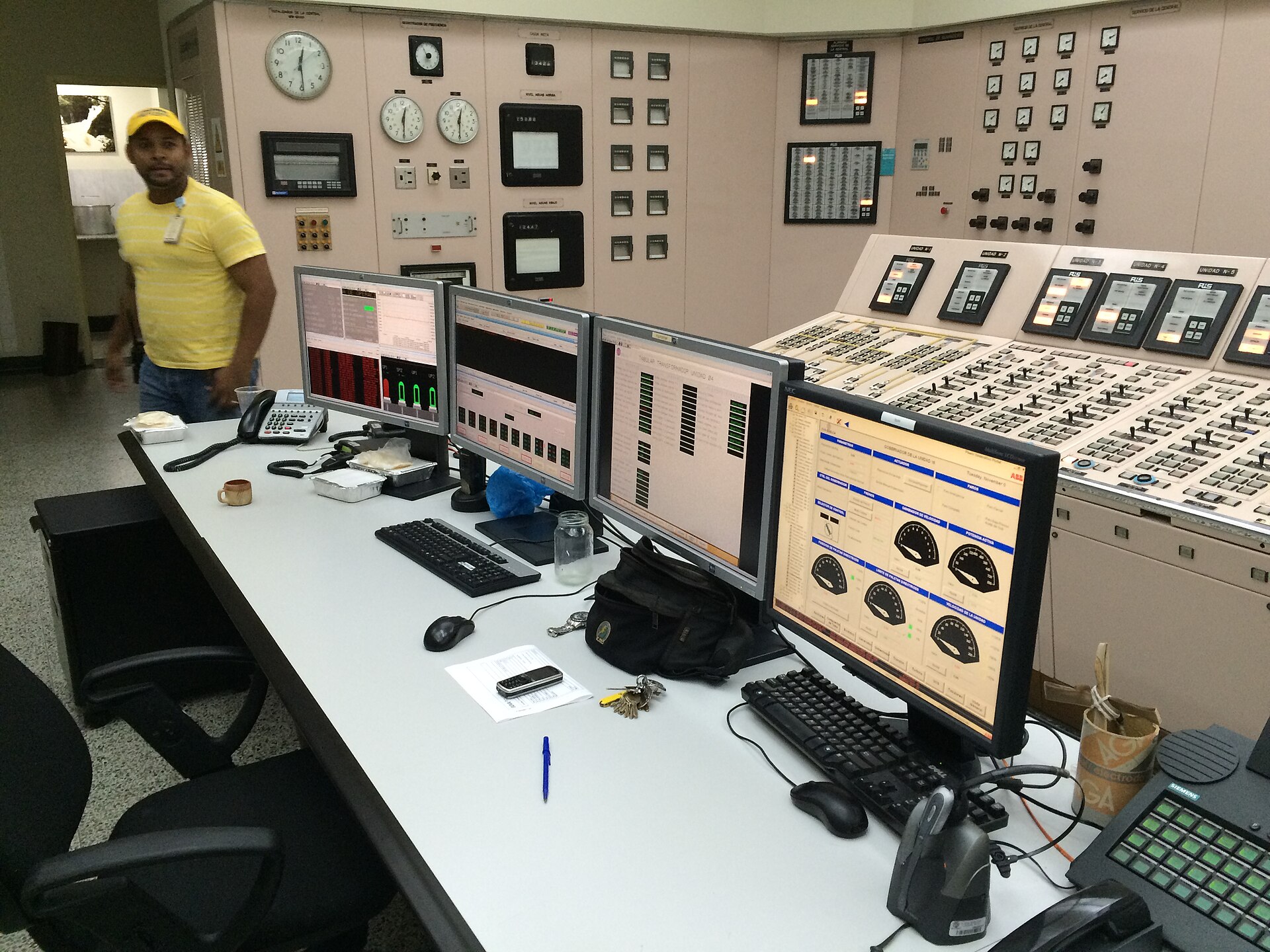 Control room with wall panels, dials and four computer monitors on a long desk, with a worker in a yellow shirt at left, at the Guri hydroelectric plant in Venezuela