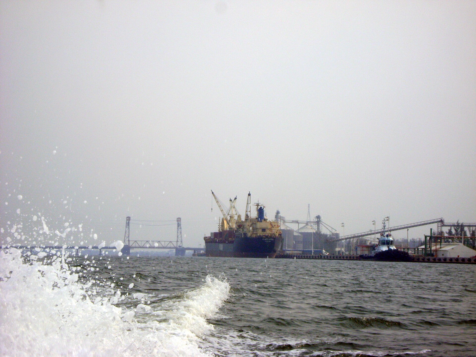 A cargo ship and a tugboat moored at an industrial river port with cranes and conveyor belts, a lift bridge in the hazy background and spray in the foreground