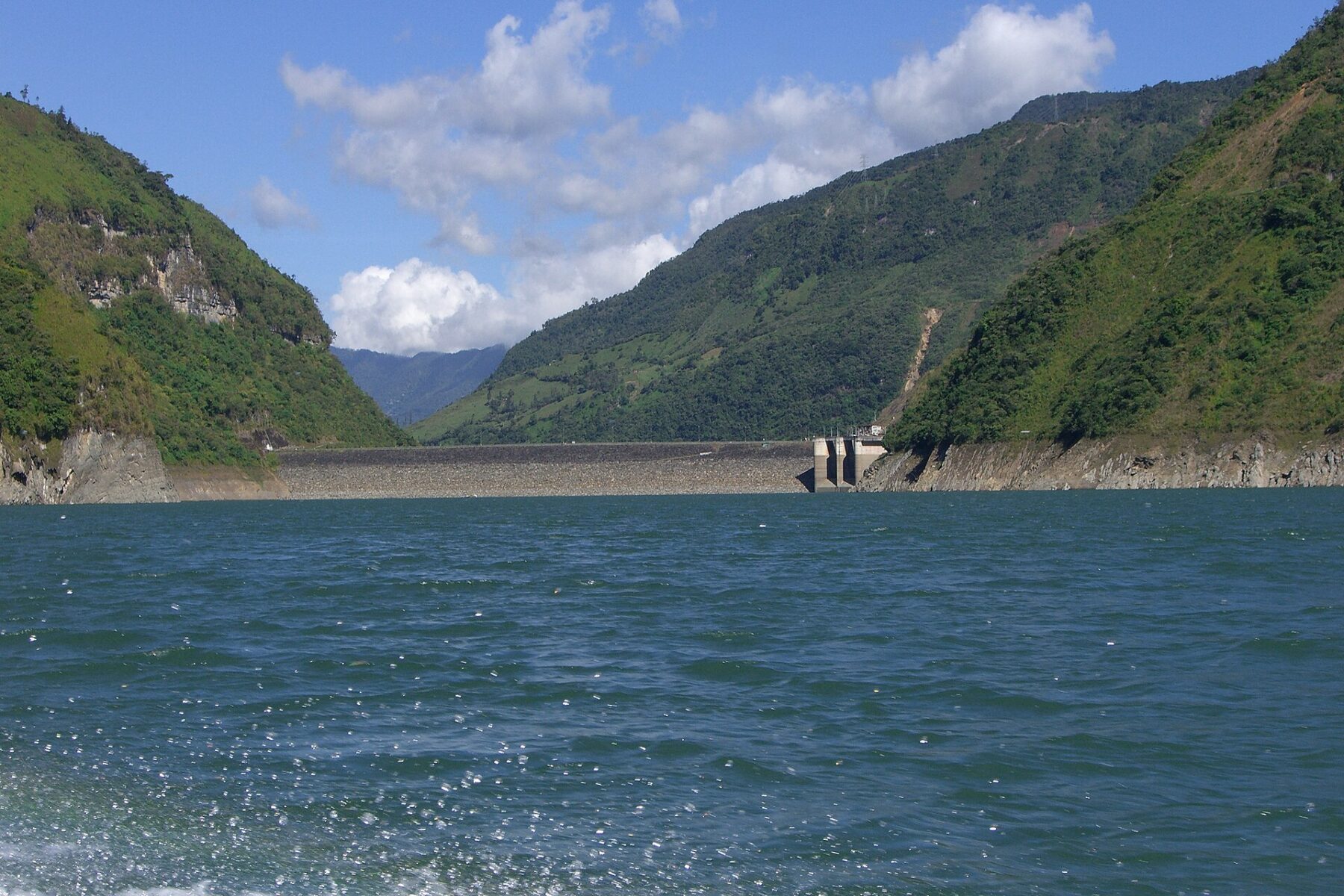 A blue-green mountain reservoir with a dam wall and intake tower between steep green hillsides under a partly cloudy sky in Colombia
