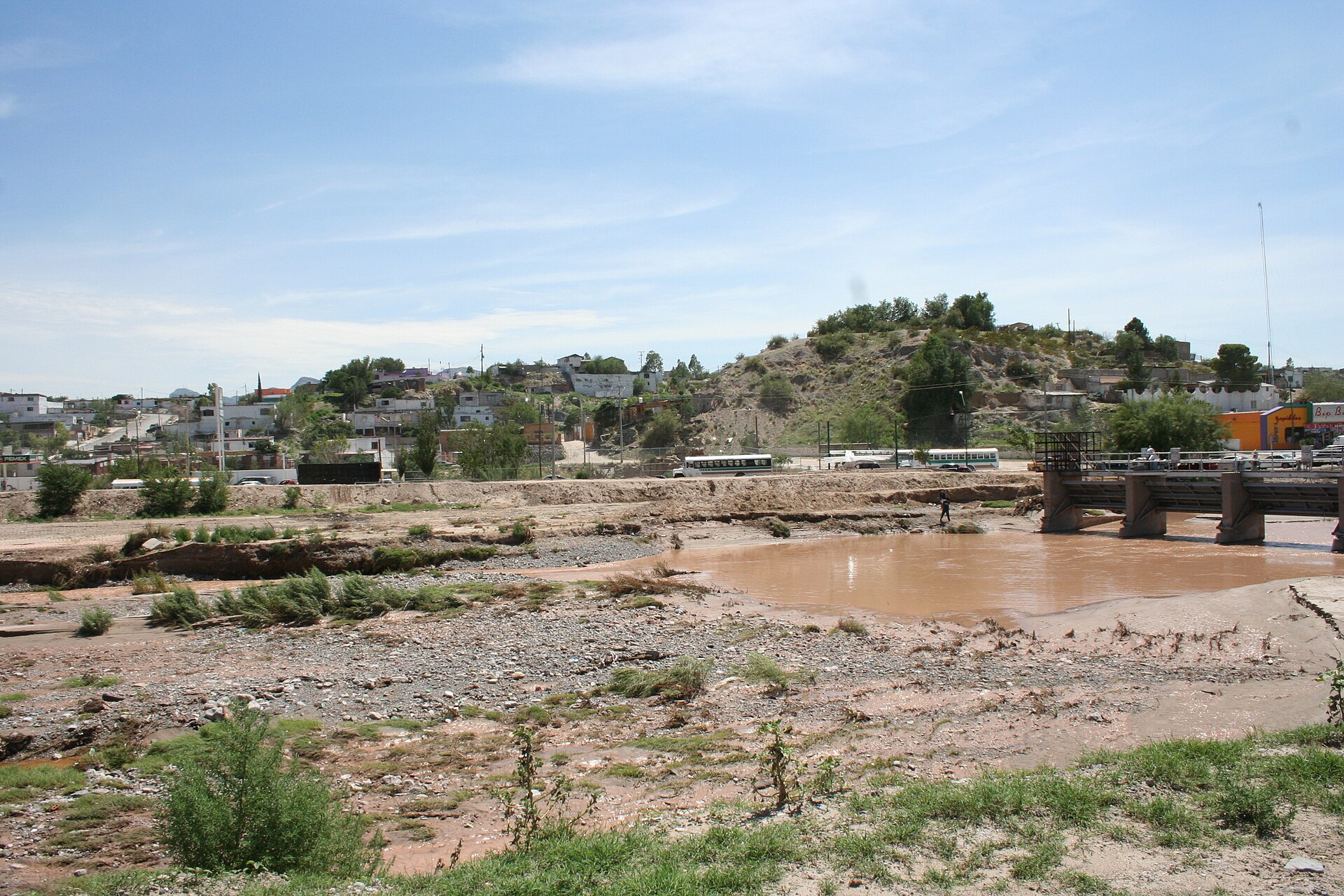 A muddy Rio Grande channel with houses and a hillside of Ciudad Juarez behind it and a small bridge at right