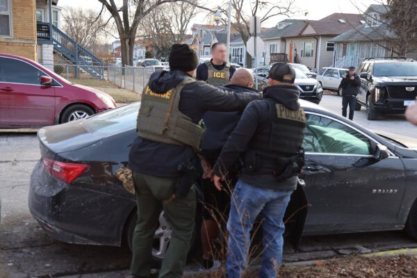 Uniformed ICE and police officers detain a man beside a dark car on a residential street, faces blurred