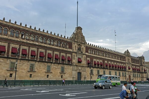 The long stone facade of the National Palace in Mexico City with red window awnings, a bus and a car passing on the street in front and pedestrians on the pavement