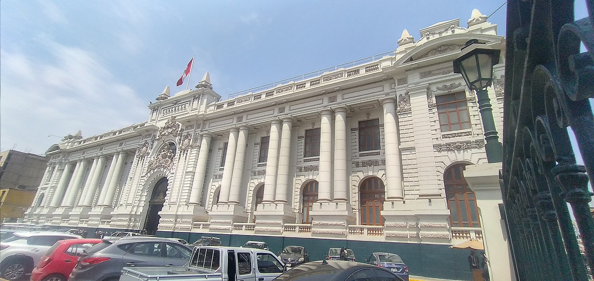 The white neoclassical facade of the Legislative Palace in Lima with columns, arched doors and a Peruvian flag, seen from the street behind an iron fence