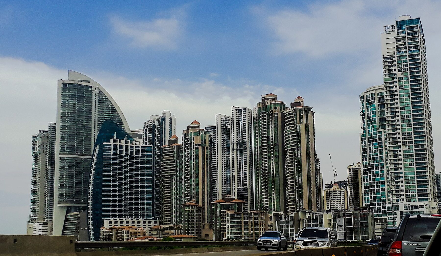 A cluster of tall glass and concrete towers in Punta Pacifica, Panama City, seen from a highway under a cloudy blue sky