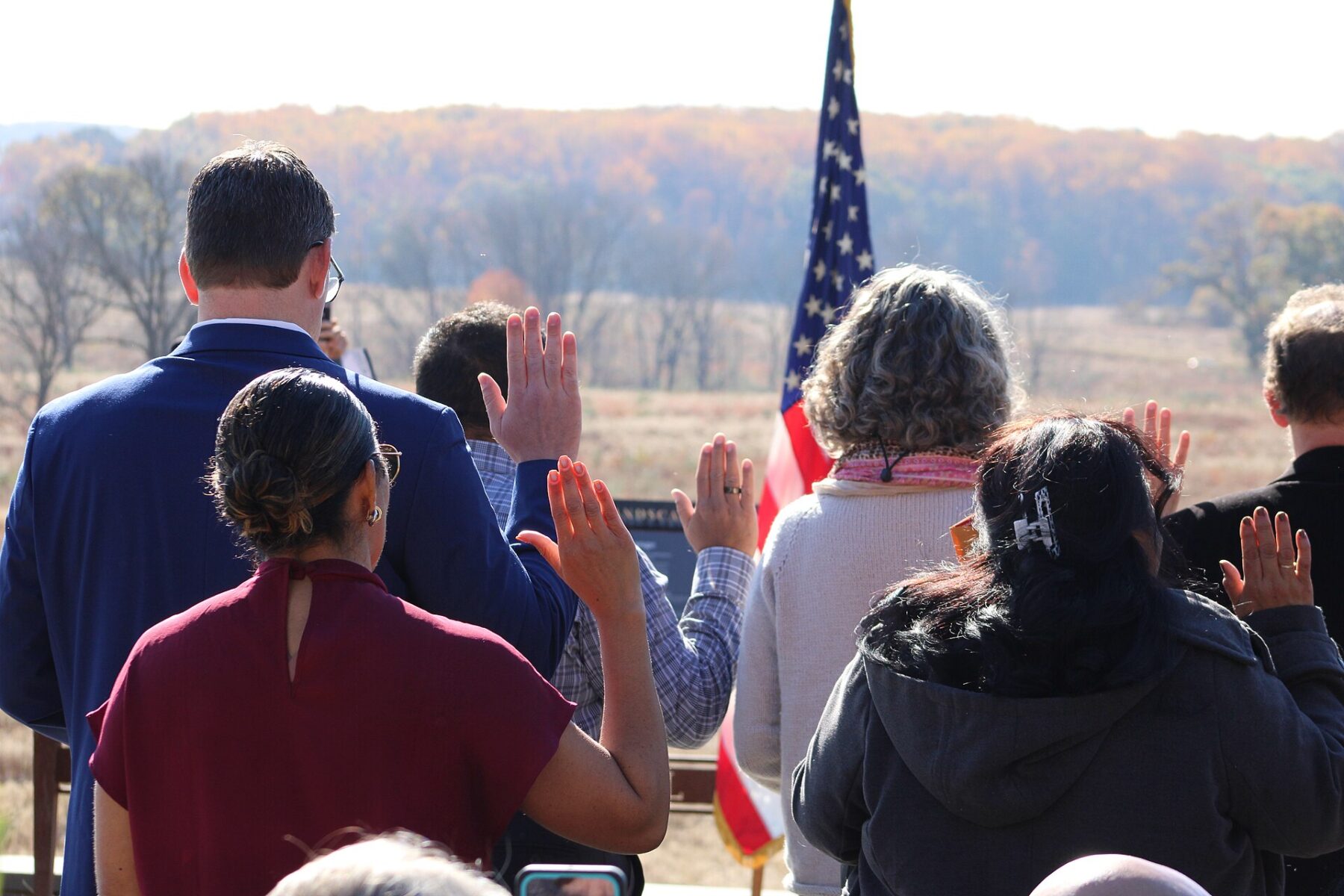 Backs of new citizens raising their right hands in front of a US flag at an outdoor oath ceremony
