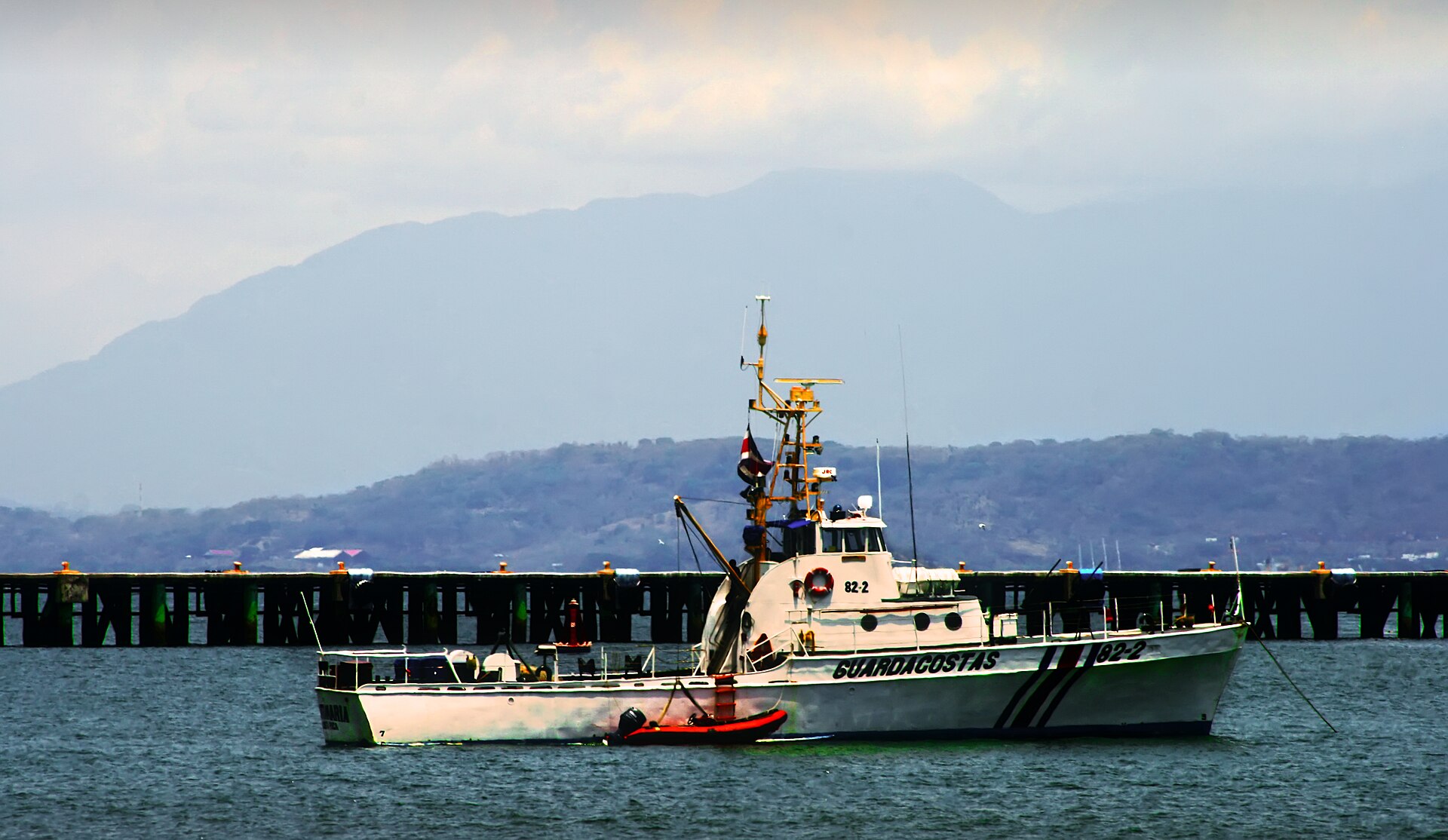 A white Guardacostas patrol boat numbered 82-2 at anchor in front of a pier and hazy mountains