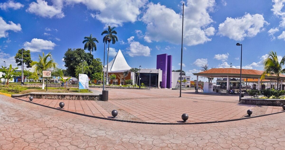 Wide view of a paved public square with palm trees, a white tent-shaped roof, a purple building and a pavilion under a blue sky in Tapachula, Mexico