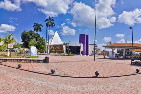 Wide view of a paved public square with palm trees, a white tent-shaped roof, a purple building and a pavilion under a blue sky in Tapachula, Mexico