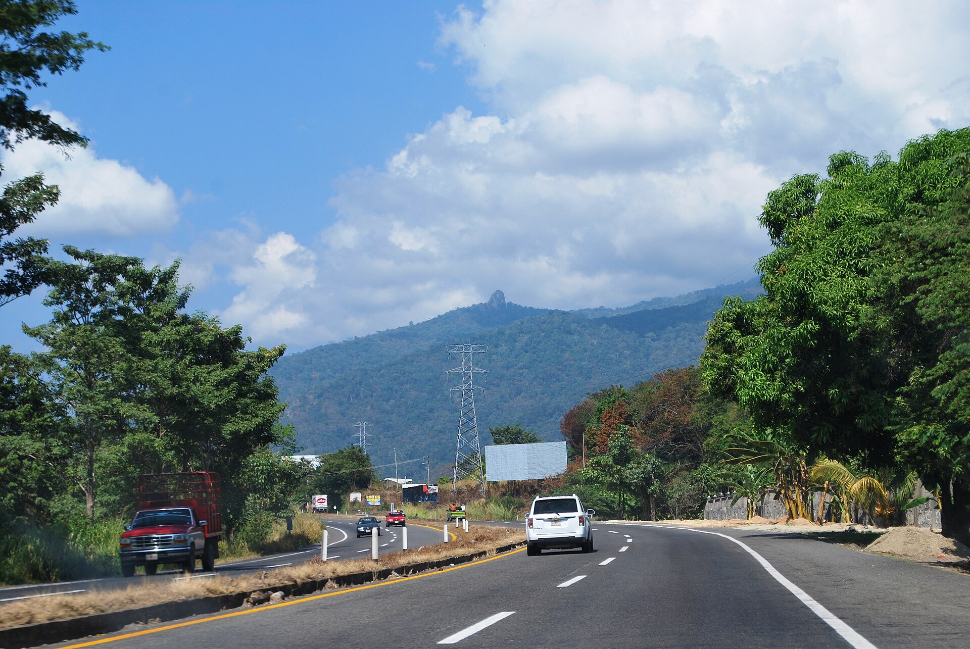 A two-lane highway with a white car and a red truck, green trees and a forested mountain with a pylon under a blue sky in southern Chiapas