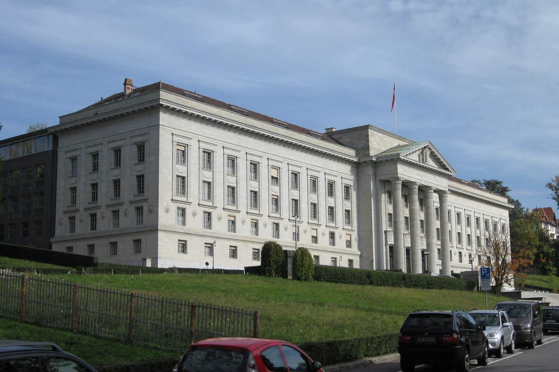 A pale stone neoclassical courthouse with a columned portico and a flag, seen from a hillside road with parked cars in the foreground