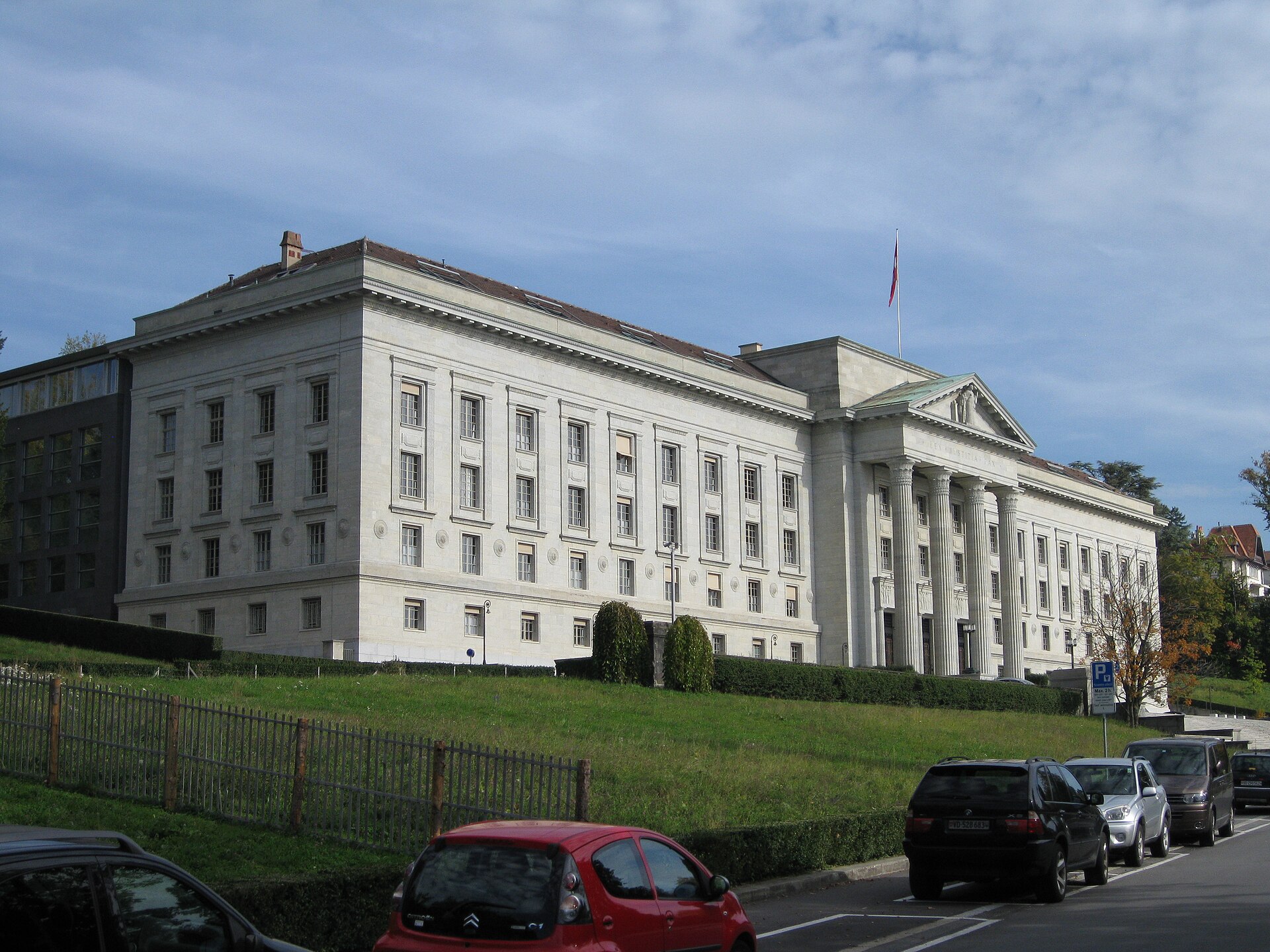 A pale stone neoclassical courthouse with a columned portico and a flag, seen from a hillside road with parked cars in the foreground
