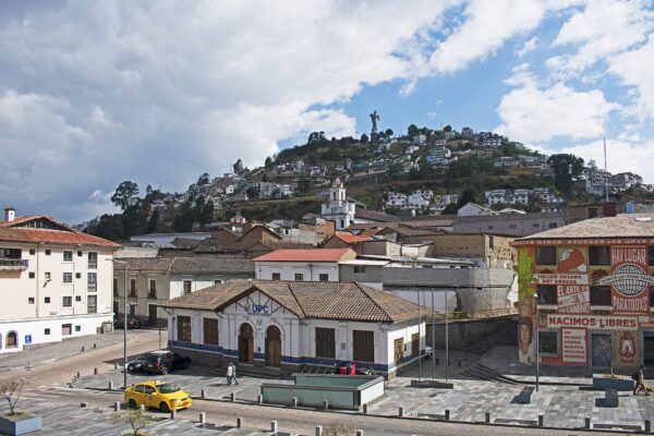 A yellow taxi on a Quito street below the hill of El Panecillo, with its winged Virgin statue above colonial rooftops under a cloudy sky