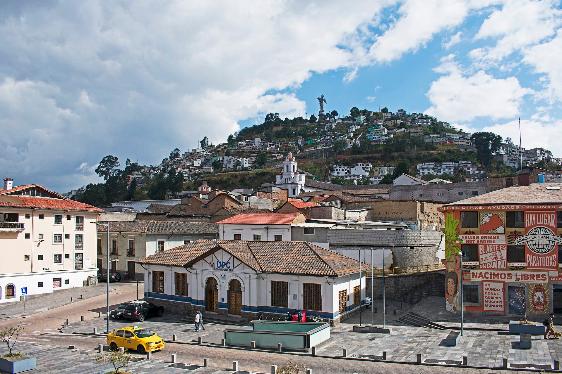 A yellow taxi on a Quito street below the hill of El Panecillo, with its winged Virgin statue above colonial rooftops under a cloudy sky