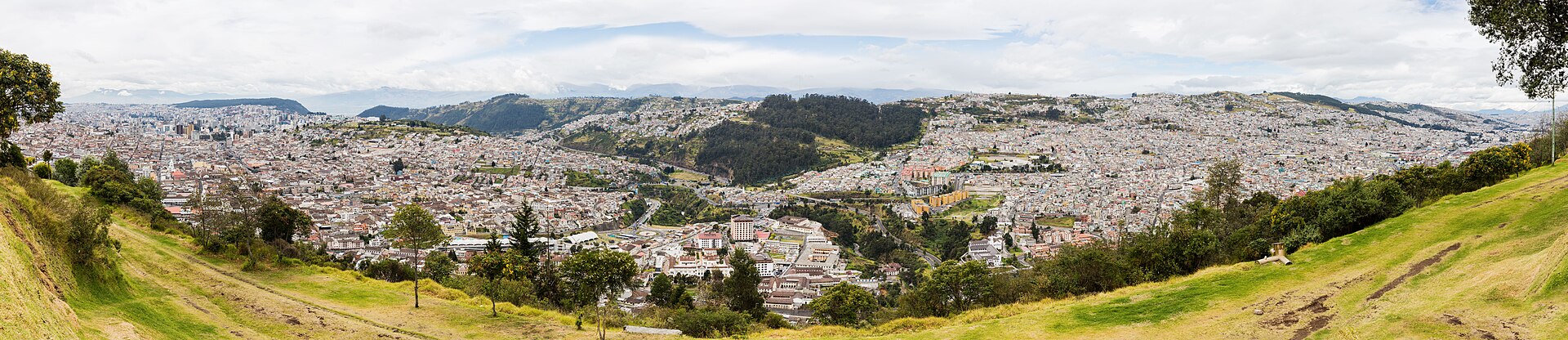 Wide panorama of Quito's rooftops and green hillsides seen from a grassy slope under a cloudy sky