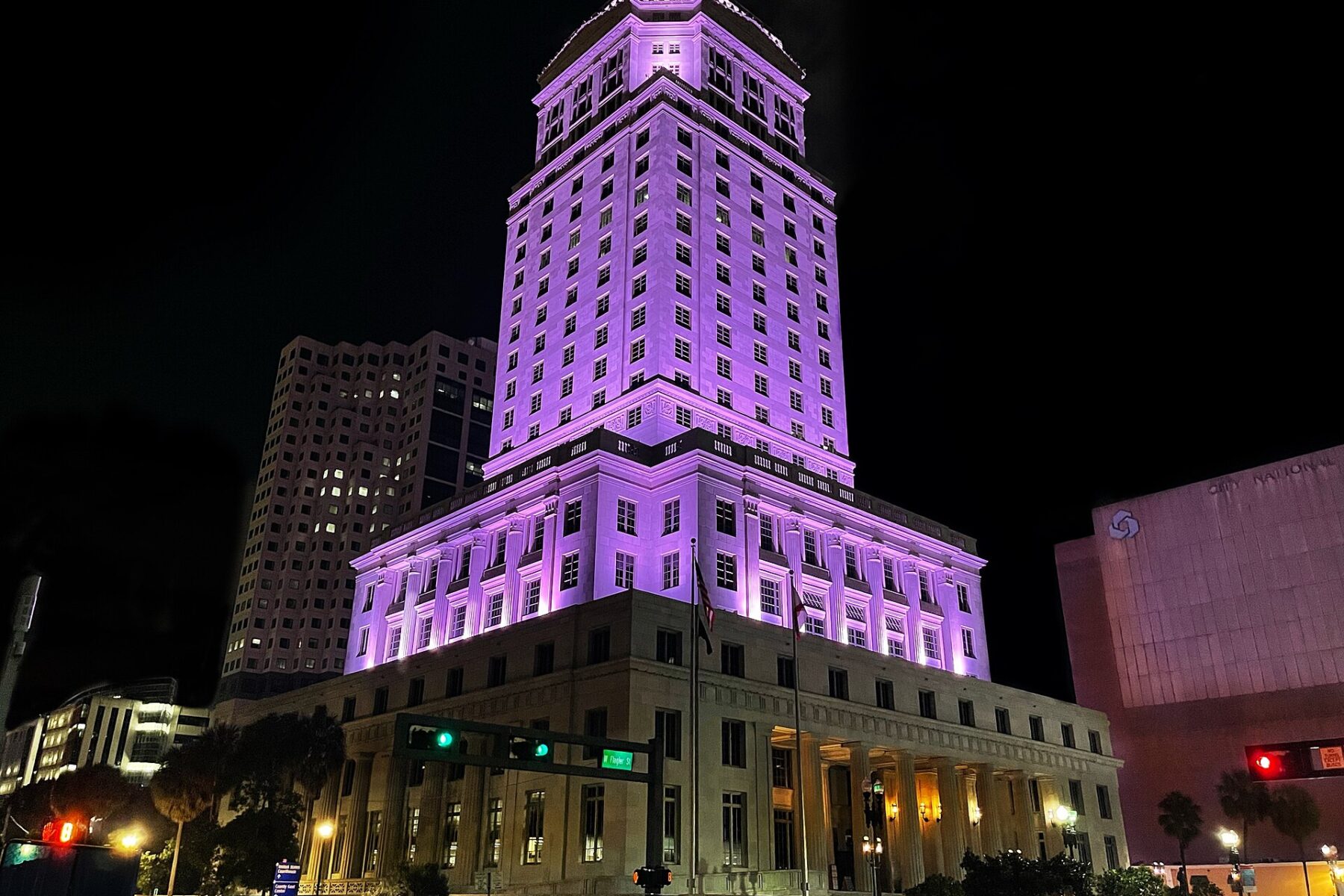 The Dade County Courthouse, a tall stone tower with a pyramid top, lit purple at night above a street with traffic lights