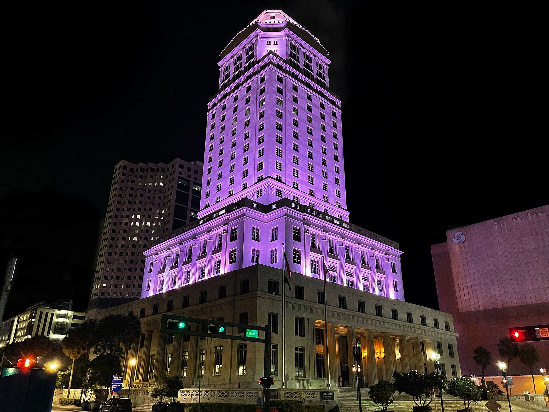 The Dade County Courthouse, a tall stone tower with a pyramid top, lit purple at night above a street with traffic lights