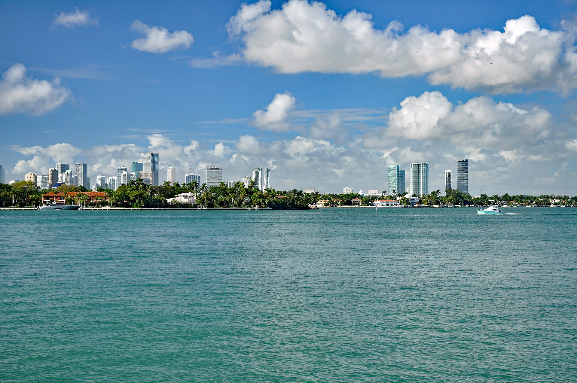 The downtown Miami skyline seen across the green water of Biscayne Bay, with palm-lined islands and a boat under a partly cloudy sky