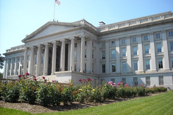 The columned stone facade of the US Treasury building in Washington, DC, with rose beds in the foreground and a flag on the roof