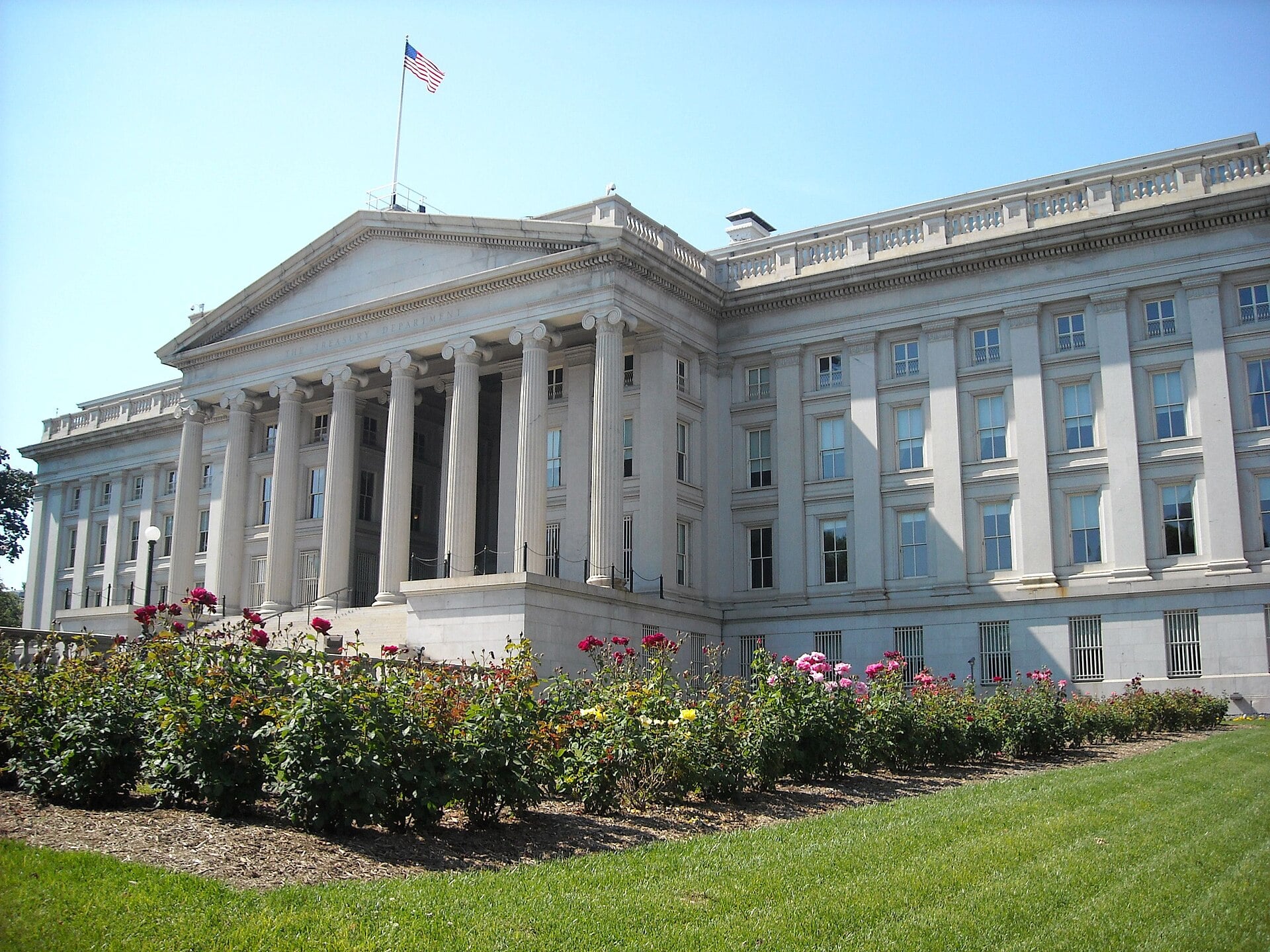 The columned stone facade of the US Treasury building in Washington, DC, with rose beds in the foreground and a flag on the roof