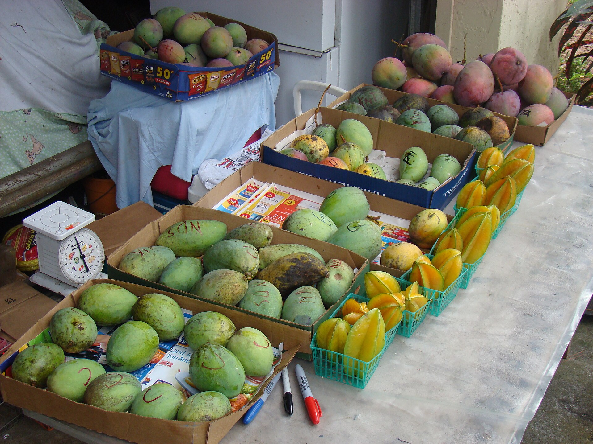 Cardboard boxes of green and red mangoes and baskets of starfruit on a table at a fruit stand, with a small kitchen scale at the left