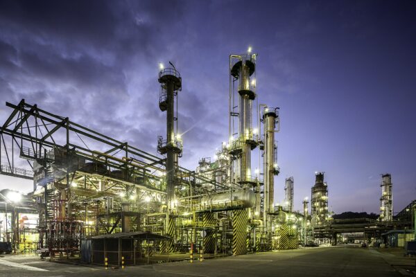 Distillation columns, pipes and walkways of an oil refinery lit up at dusk under a dark blue cloudy sky