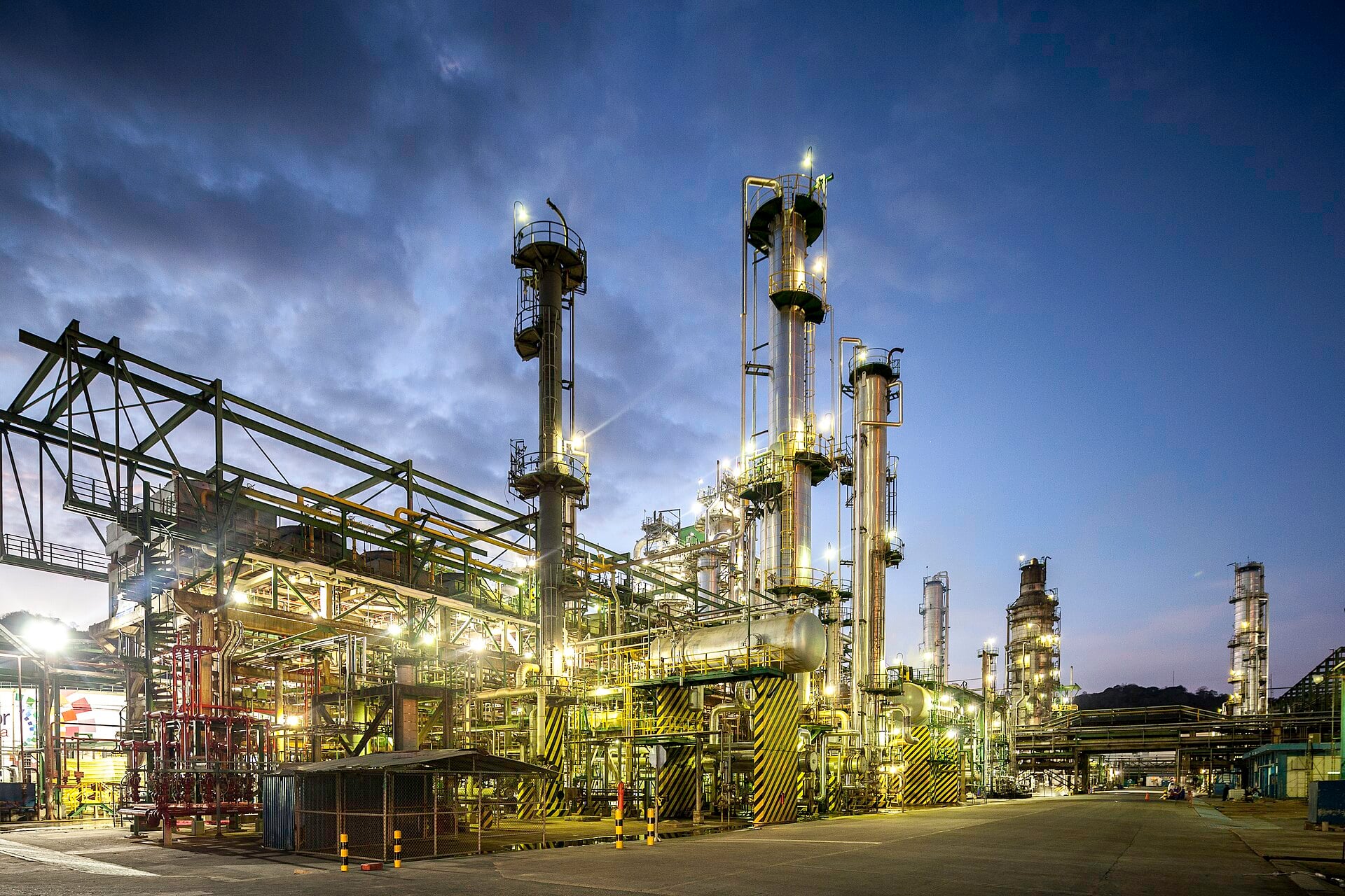 Distillation columns, pipes and walkways of an oil refinery lit up at dusk under a dark blue cloudy sky