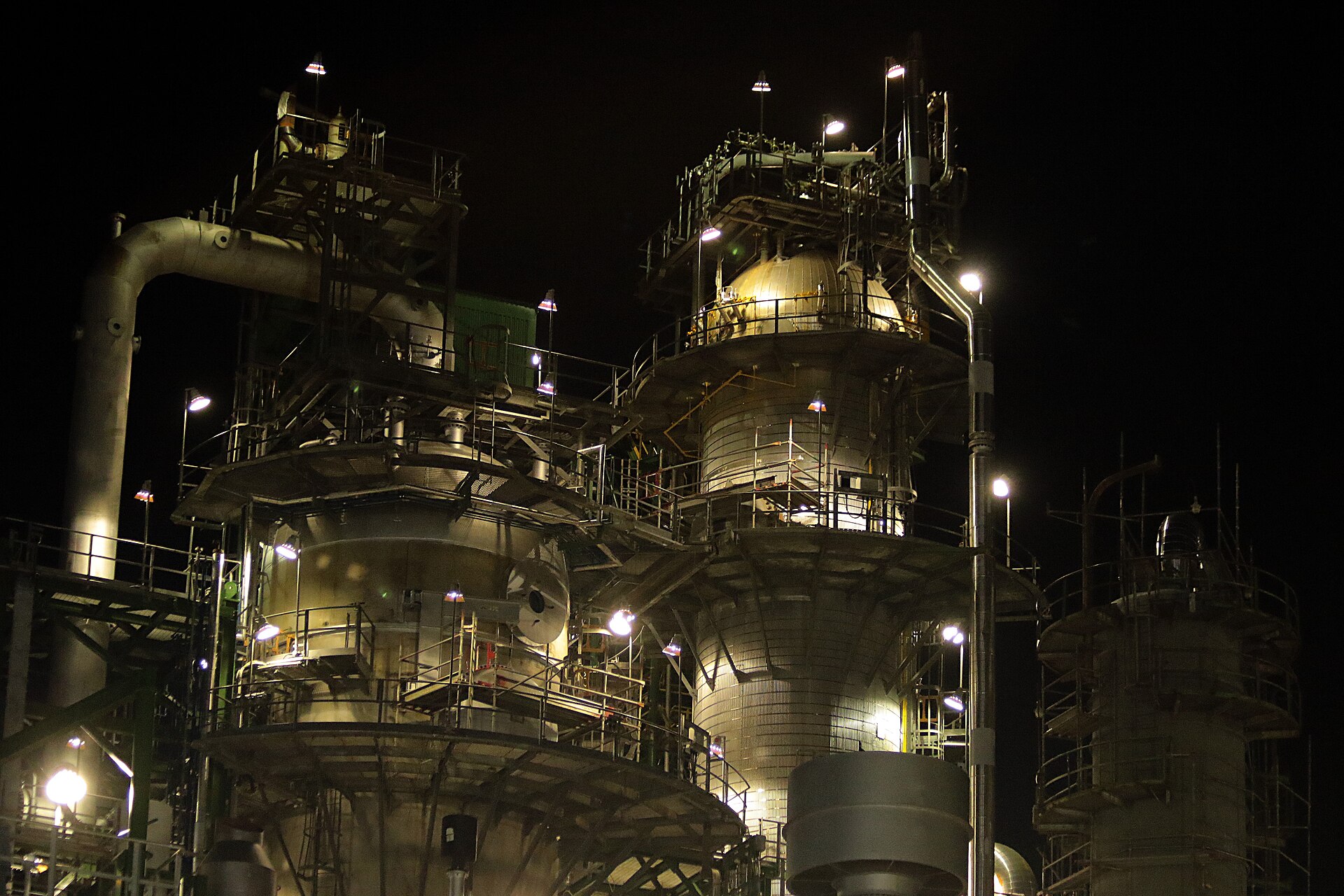 Close view of refinery towers, pipes and steel walkways lit by floodlights against a black night sky
