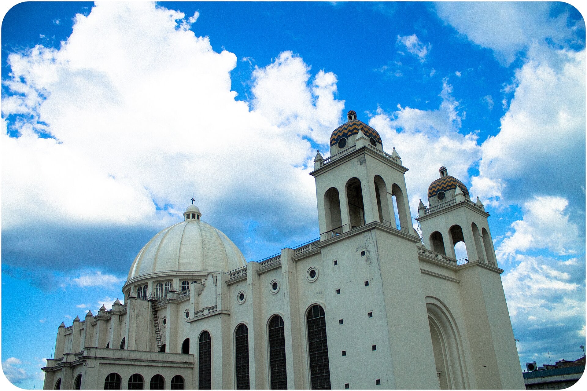 The white domed Metropolitan Cathedral of San Salvador with two bell towers under a blue sky and large white clouds