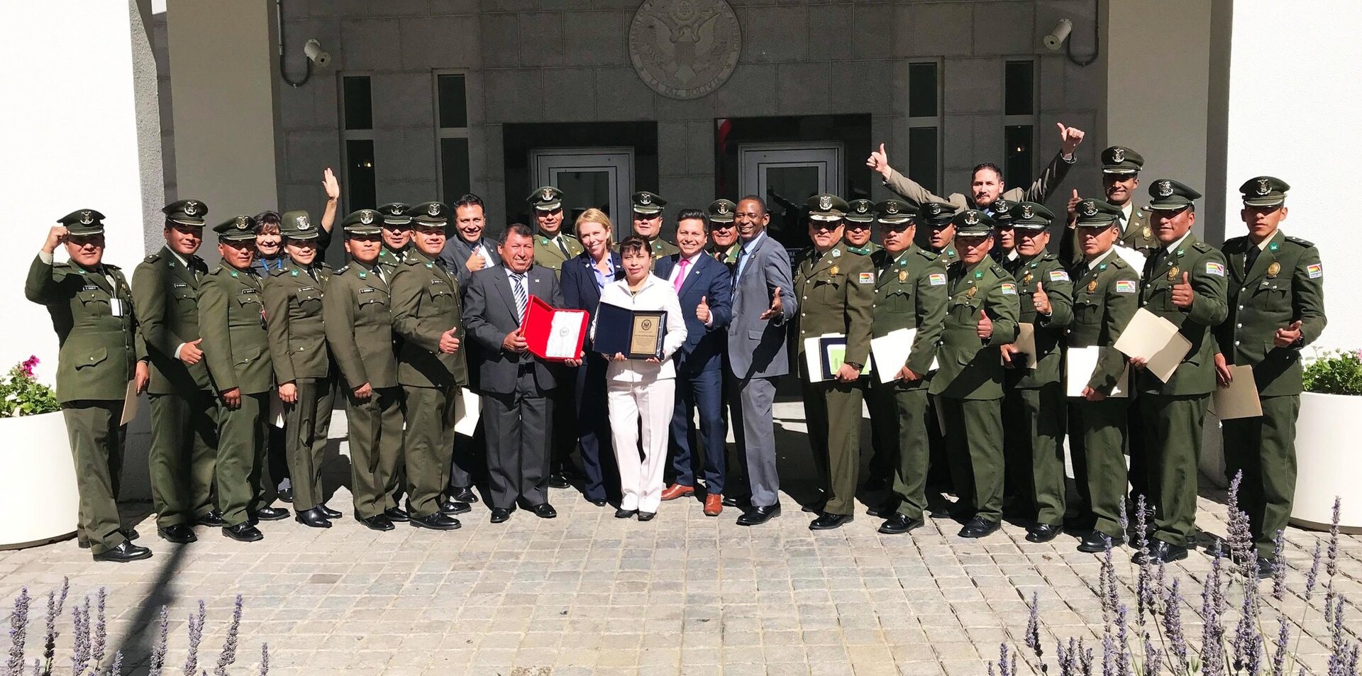 Bolivian police officers in olive-green uniforms and US Embassy staff in suits posing together outside the embassy entrance, some giving thumbs up