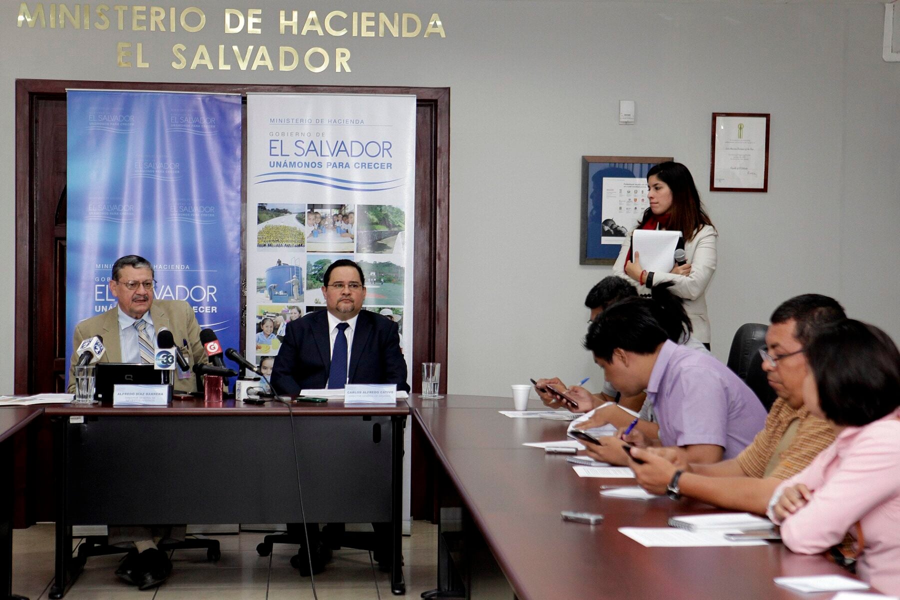 Two officials at a table in front of Ministerio de Hacienda El Salvador banners, with reporters taking notes in a meeting room