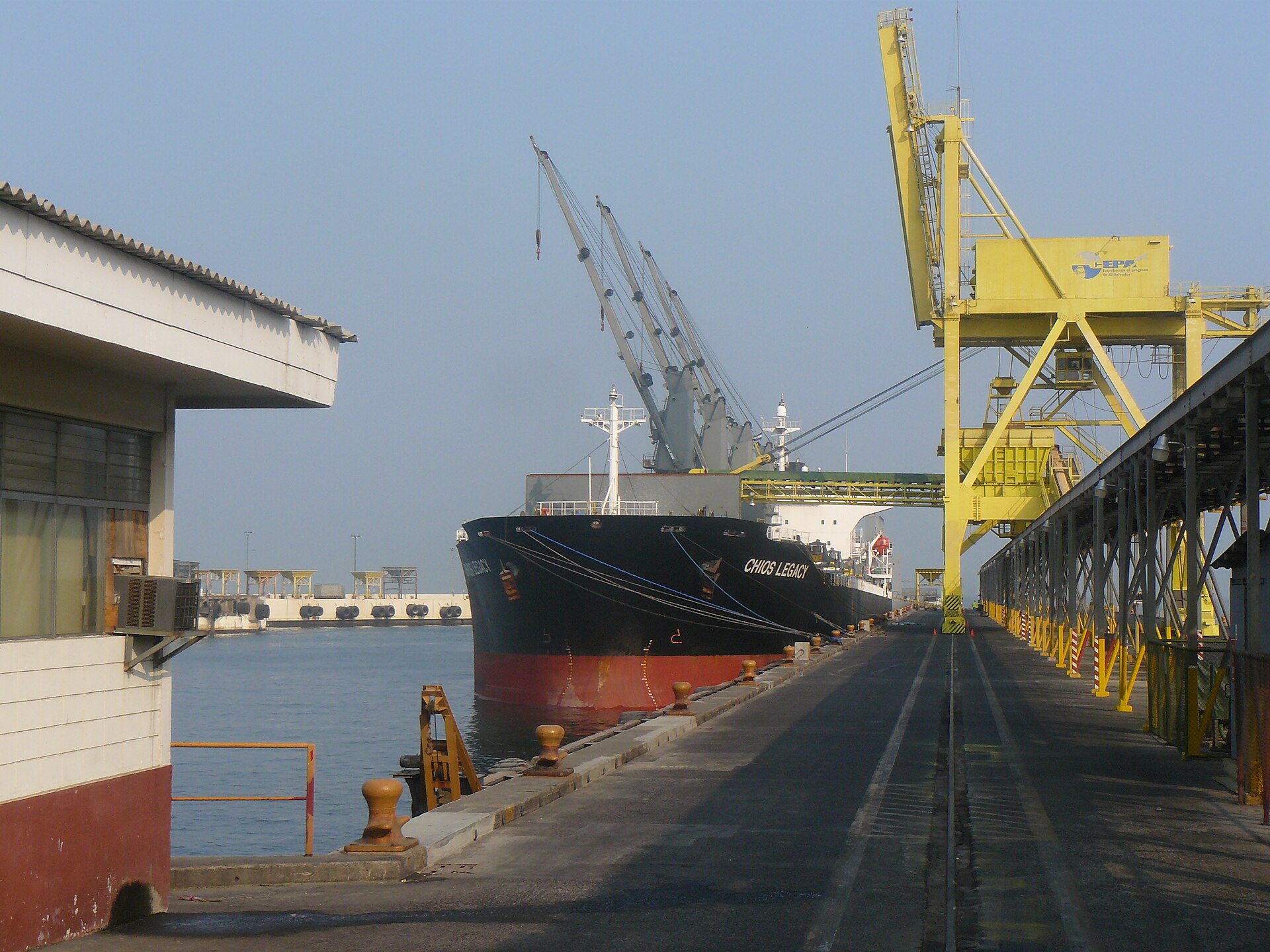 A black bulk carrier with red hull moored at a concrete quay in the port of Acajutla, with a yellow gantry crane and cargo cranes under a hazy sky