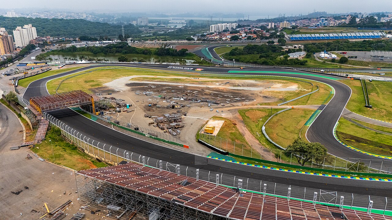 Aerial view of the Interlagos motor-racing circuit in São Paulo, with a curved asphalt track, grass infield, temporary stands in the foreground and apartment blocks beyond.