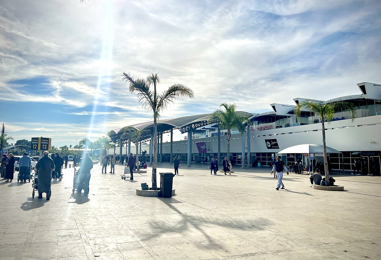 Crowds of travelers and luggage trolleys on the sunlit forecourt of Mohammed V International Airport in Casablanca, with palm trees and the arrivals building behind.